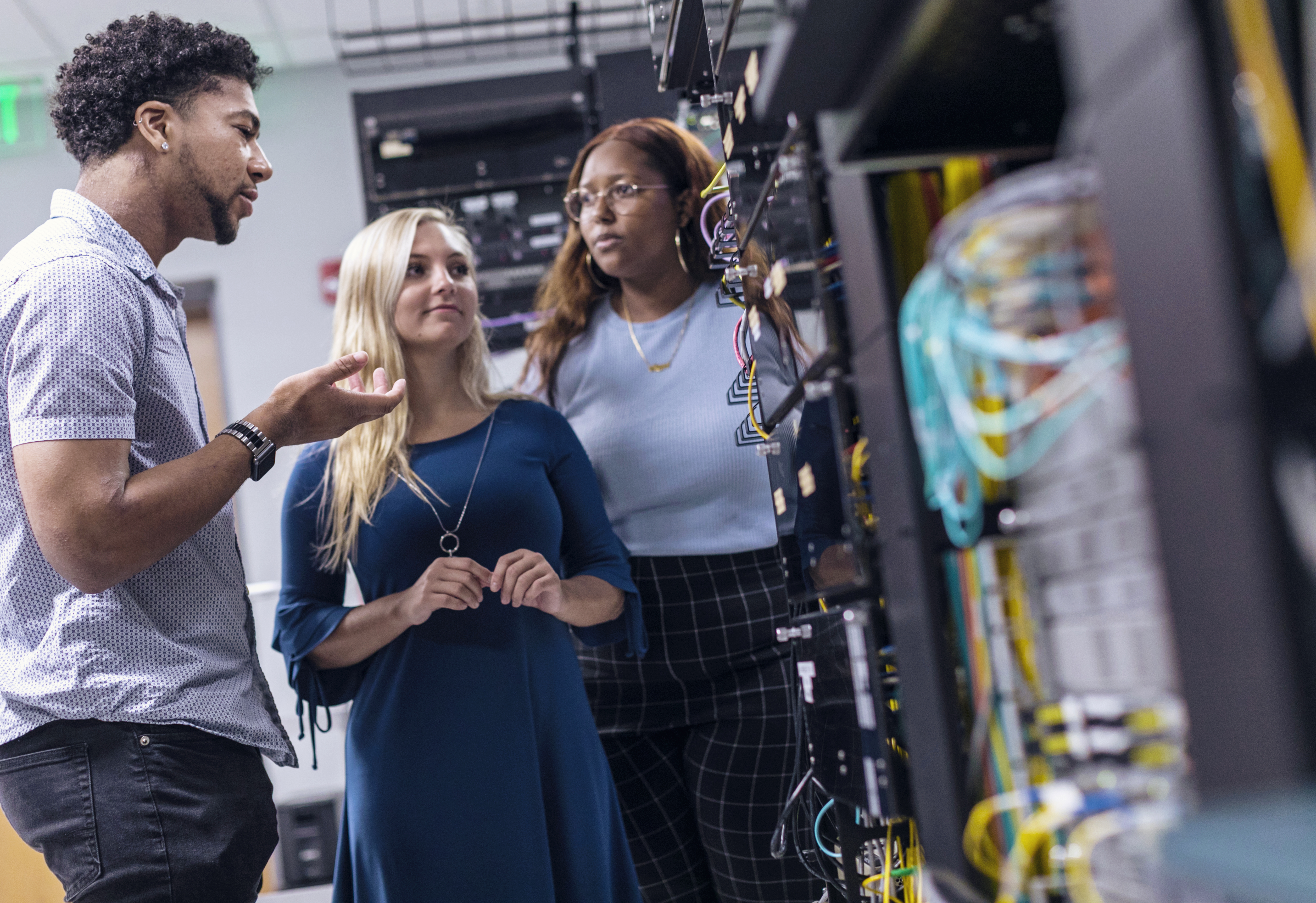 A group of three students are standing around electrical equipment in the cyber security lab