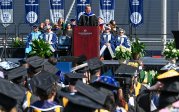 A man in academic regalia speaks to students before students wearing robes and mortarboards.